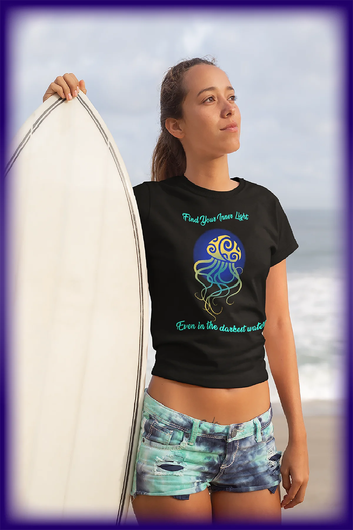 Young woman holding surfboard at beach wearing black t-shirt with jellyfish and inspirational quote